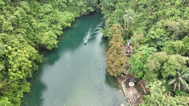 Aerial view of a boat traveling along the green river surrounded by dense tropical forest and canyon walls in Pangandaran Regency, West Java, Indonesia.