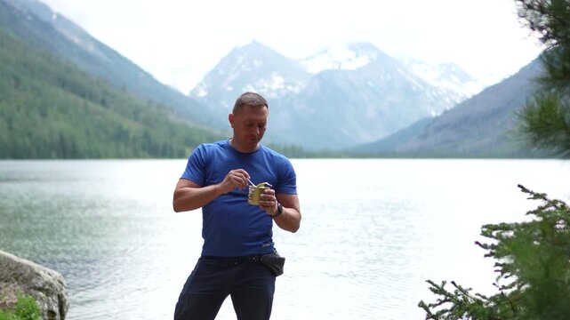 Medium portrait of an athletic male hiker in a blue t-shirt standing on a lake shore, eating canned food and looking at snow-capped mountain peaks.