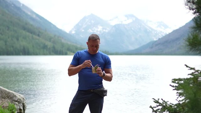 Medium portrait of an athletic male hiker in a blue t-shirt standing on a lake shore, eating canned food and looking at snow-capped mountain peaks.