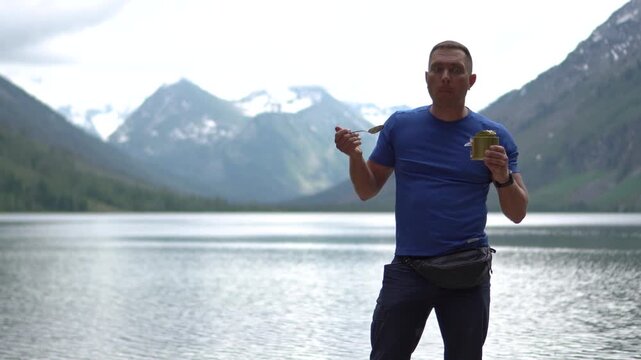 Medium portrait of an athletic male hiker in a blue t-shirt standing on a lake shore, eating canned food and looking at snow-capped mountain peaks.