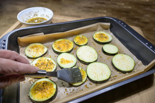 Hand with a silicone brush spreading seasoned oil on eggplant slices on a baking sheet, preparing aromatic roasted vegetables, selected focus