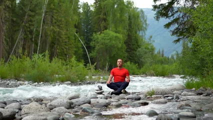 Fototapeten Lotusblume A man travels through the Altai Mountains. A man in red meditates and practices breathing exercises on the banks of a mighty mountain river. A man sits in the lotus position on the riverbank, feeling   © OlTarakanov