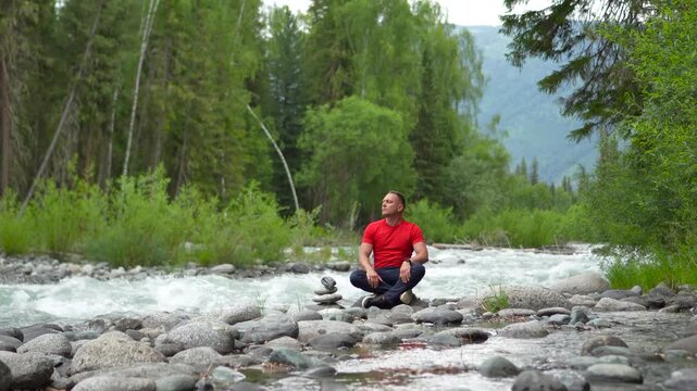 A man travels through the Altai Mountains. A man in red meditates and practices breathing exercises on the banks of a mighty mountain river. A man sits in the lotus position on the riverbank, feeling 