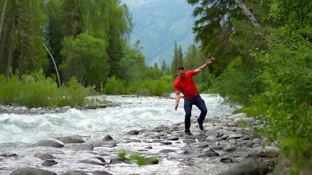 A man is traveling through the Altai Mountains. A man in red is standing on the bank of a powerful mountain river, trying to hold on to the rocks.