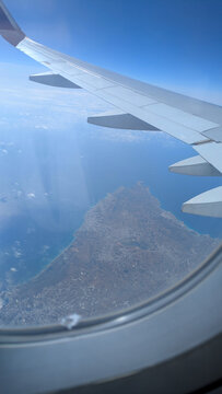 Aerial view from an airplane window showing a wing over a coastal island and ocean under clear blue skies.
