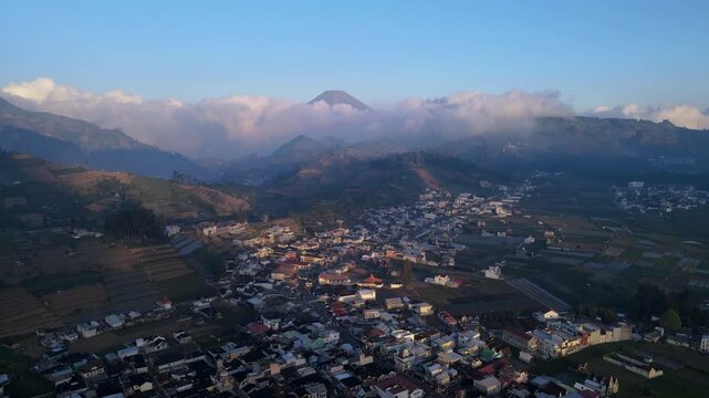 Aerial view of Bukit Skoter overlooking the town of Kecamatan Batur with terraced fields and distant mountains under a clear sky in Kecamatan Batur, Jawa Tengah, Indonesia.