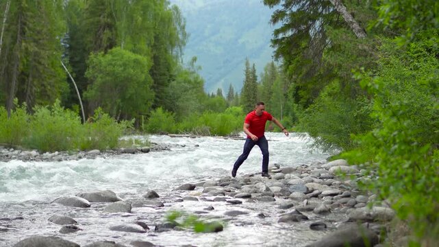A man travels through the Altai Mountains. A man in red on the bank of a powerful mountain river hops over rocks to keep his shoes dry. A man on the riverbank in Altai.