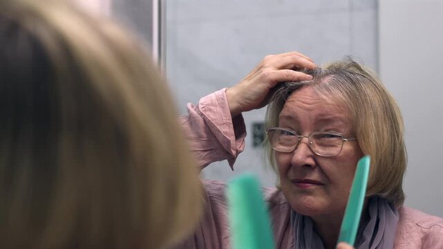 Mature woman looking in a bathroom mirror and checking her hair with a comb. Hair care, gray hair and personal grooming scene. Gray hair care and personal grooming scene.