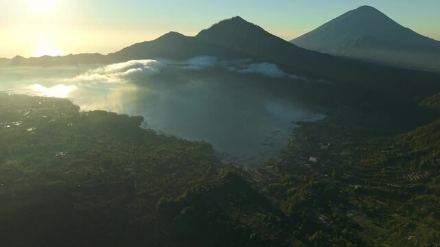 Aerial view of Lake Batur and Mount Abang at sunrise with golden light reflecting on the water and clouds over the volcanic landscape in Kintamani, Bali, Indonesia.
