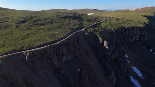 Aerial drone footage captures vehicle traversing dramatic Beartooth Highway in Montana, USA, winding along steep rocky cliffs with lush green alpine meadows and patches of snow under clear blue skies.
