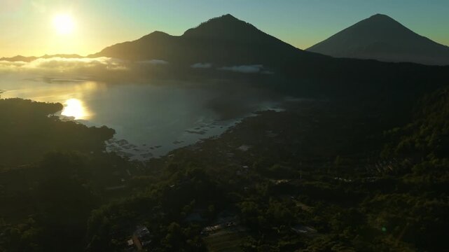 Aerial view of Lake Batur and Mount Abang at sunrise with golden light reflecting on the water surface and lush volcanic hills in Kintamani, Bali, Indonesia.