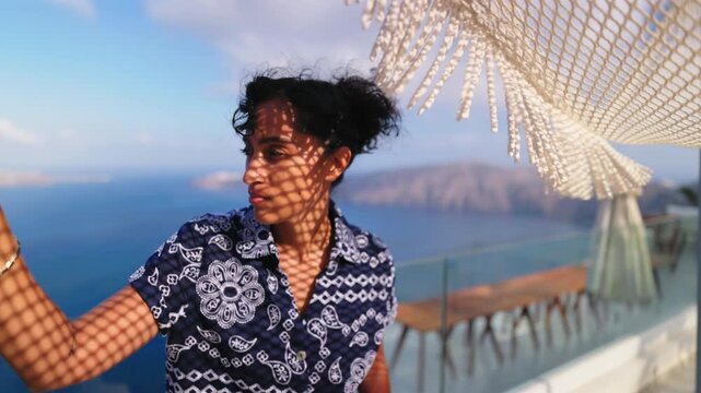 Young Indian woman with curly hair stands under woven macrame canopy on terrace overlooking caldera in Santorini Greece. Golden hour light creates pattern shadows on her face and patterned shirt.