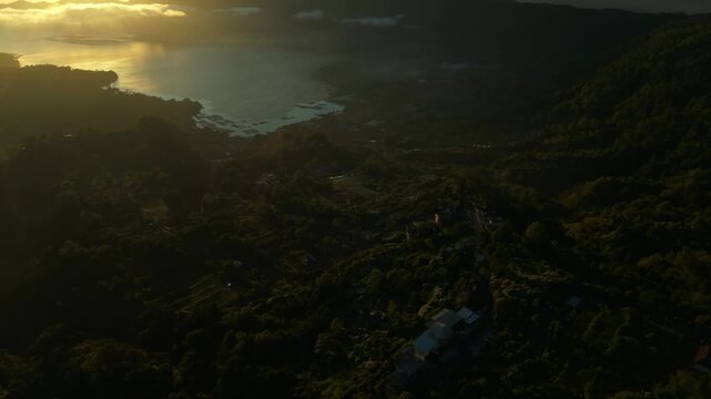 Aerial view of Lake Batur and Mount Abang at sunrise with golden light reflecting on the water and lush volcanic slopes in Kintamani, Bali, Indonesia.