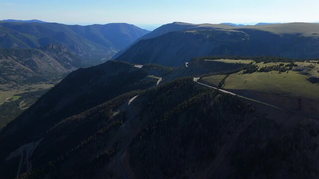 Aerial of Beartooth Highway in Montana, USA featuring dramatic switchbacks winding through forested mountainside. Drone footage captures scenic valley views with layered peaks and alpine meadows.