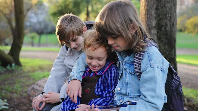 Three young children sit on a fallen log in a sunny park, gathered around a smartphone. Older siblings share device with toddler sibling in autumn setting with green grass and trees.