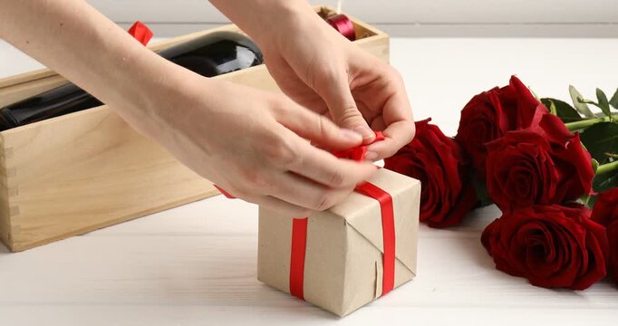 Woman with gift box, bottle of wine and red roses at white wooden table, closeup