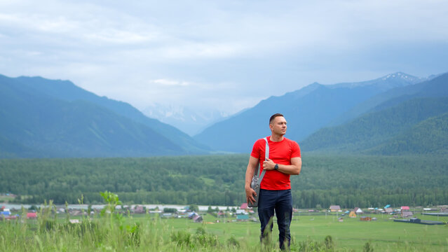 A man travels through the Altai Mountains. A man in red poses against a backdrop of distant mountain peaks, Mount Belukha hidden by clouds. A male traveler in bright clothing poses for a photo.
