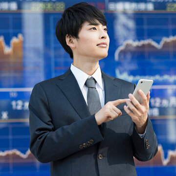 株価ボード前でスマートフォンを操作する20代男性ビジネスマン