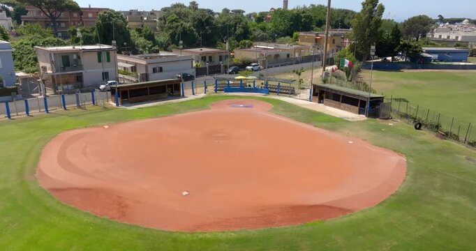 An elevated drone shot captures a scenic baseball diamond, emphasizing the large red clay infield, surrounding green grass, team dugouts, perimeter fences, and local homes under a bright and clear sky