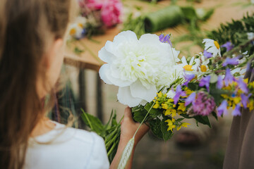 Naklejka premium Young girl holding a bouquet of colorful flowers including white peony and purple blooms, with a blurred background of additional floral arrangements on a wooden table