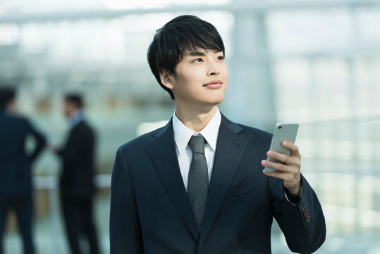 オフィスビルのロビーでスマートフォンを操作する20代男性ビジネスマン