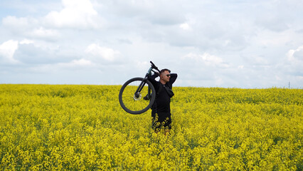 Fototapeta premium Moments of cycling. A man poses with his bicycle in a rapeseed field; a male athlete holds his bicycle behind his head, enjoying the views of the yellow rapeseed field.