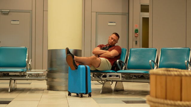 Air travel. A tired man waits for his delayed flight at the airport. The man rests his feet on his suitcase and dozes off while waiting.
