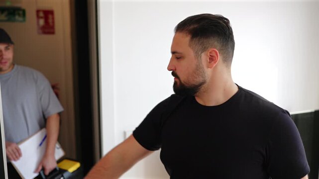 Professional service technician wearing cap and gray shirt arrives at modern apartment doorway holding clipboard while homeowner in black t-shirt opens door and greets him for scheduled maintenance.