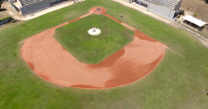 Aerial drone shot of an empty baseball field diamond during a sunny morning, highlighting the red clay infield dirt, green grass turf, pitcher's mound, and clear white baseline markings clearly.