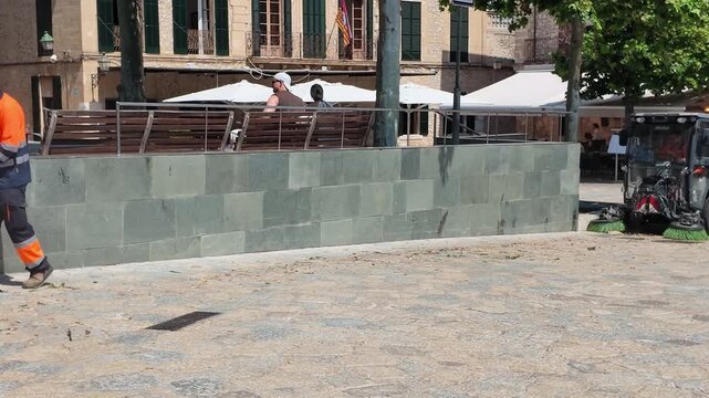 Pollensa Mallorca, Spain. 01.06.2026. Video. A compact street sweeper vehicle with rotating brushes cleans debris from a stone-paved plaza in front of historic buildings and outdoor seating area