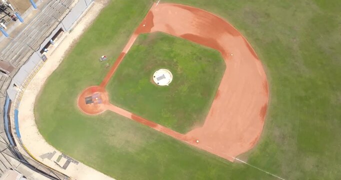 Overhead top-down aerial drone view of a beautiful baseball diamond, featuring its green grass infield and outfield, the red dirt base path, a pitcher's mound, and curved stadium bleachers.