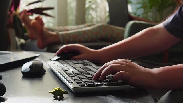 Boy&rsquo;s hands actively controlling a computer game using a keyboard and mouse on a dark desk. In the background, a girl in colorful leggings lies on a chair, gently moving her toes while using a laptop.