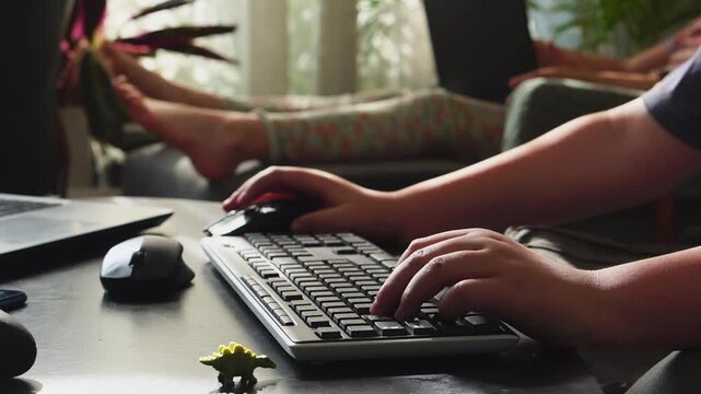 Boy playing computer game using keyboard and mouse with toy dinosaur on desk, girl with laptop in background