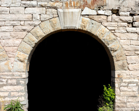 weathered stone archway set into a rough, multi-toned masonry wall. The dark opening at the center suggests it may be part of an old building or tunnel entrance
