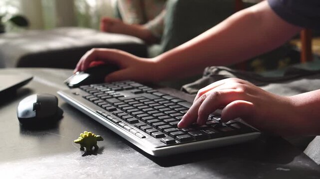 Сhild&rsquo;s hands typing on a keyboard with a small toy dinosaur nearby, while another child sits in the background. 