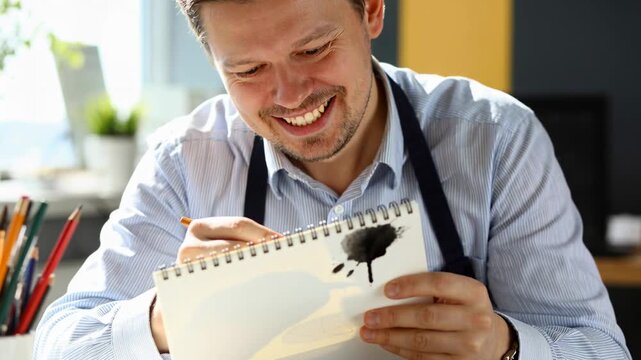 Noticing ink blot, artist tapping brush at desk with sketchbook, laughing in shirt and apron