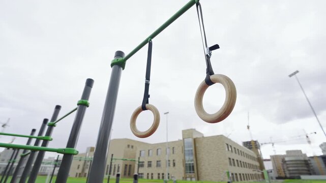 Wooden gymnastic rings hang from metal bar at outdoor workout station. Modern school building and construction cranes fill background