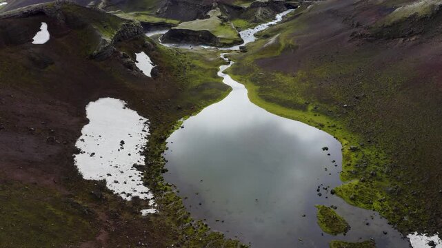 Aerial view of a winding river flowing into a glacial lake surrounded by mossy volcanic hills and patches of snow in Iceland.