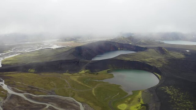 Aerial view of volcanic craters with dark soil and green mossy patches surrounded by glacial rivers under a misty sky in Iceland.