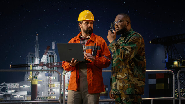Military man on drilling rig speaks into walkie talkie, enforcing maritime blockade protocol, Soldier on offshore platform talks in handheld radio to receive update on war causing oil crisis, camera.
