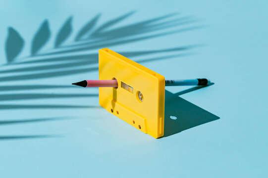 Creative retro summer music concept featuring a yellow audio cassette tape with a pencil inserted for manual rewinding and palm shadow on blue background. Nostalgic analog technology.