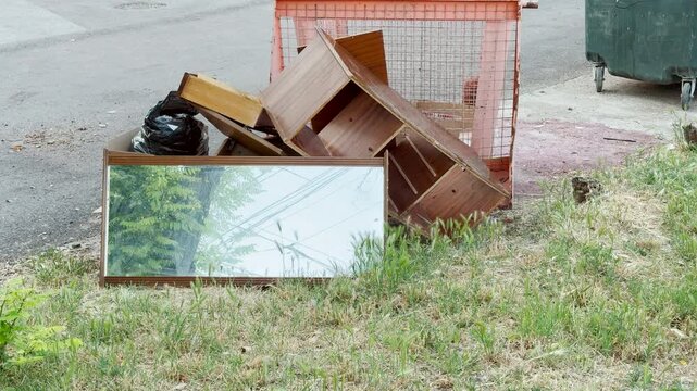 Old wooden furniture and a large mirror sit abandoned on a grassy roadside. This messy pile of discarded items brings a sad, nostalgic feeling of neglect to the neighborhood.