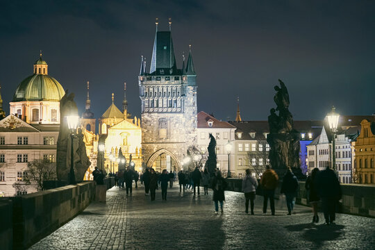 Prague, Czechia - 03 November 2021: View of Charles Bridge at night with historic statues, illuminated gothic towers, and people walking along the cobblestone path.