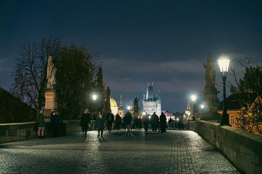 Prague, Czechia - 03 November 2021: View of Charles Bridge at night with illuminated street lamps, stone statues, and people walking along the cobblestone path towards the bridge tower.