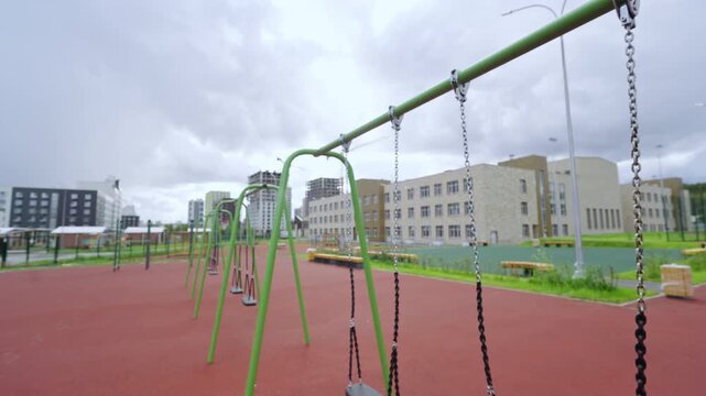 Row of swings stands on rubber surface of school playground. Contemporary educational building and construction cranes fill background