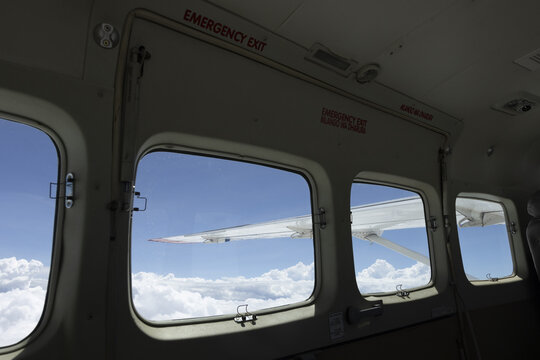 Nairobi, Kenya - 31 May 2026: View of the wing of a small airplane through cabin windows with an emergency exit sign above, flying over white clouds under a clear blue sky.