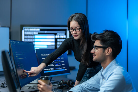 Asian woman in glasses points at a monitor of code to explain it to a seated male colleague holding coffee, collaborating in a dark blue office.