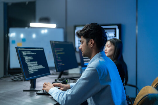 South Asian man in glasses works at his monitor of code in profile while a female colleague codes beside him in a dark blue development office.
