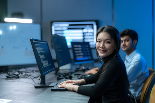 Asian woman in a black top smiles at the camera while typing at a coding workstation, with a male colleague working beside her at night.