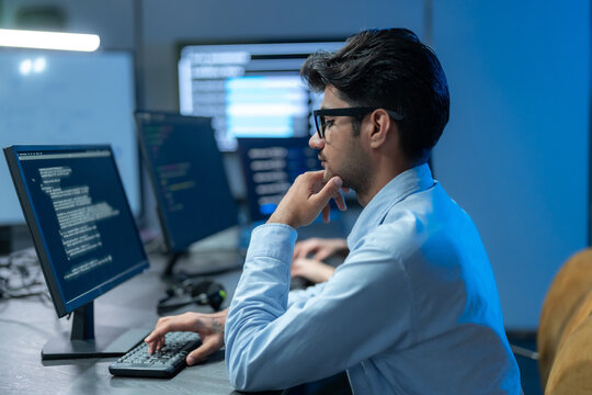 South Asian man in glasses rests his hand on his chin in thought while coding at a multi-monitor setup in a dark blue tech office at night.